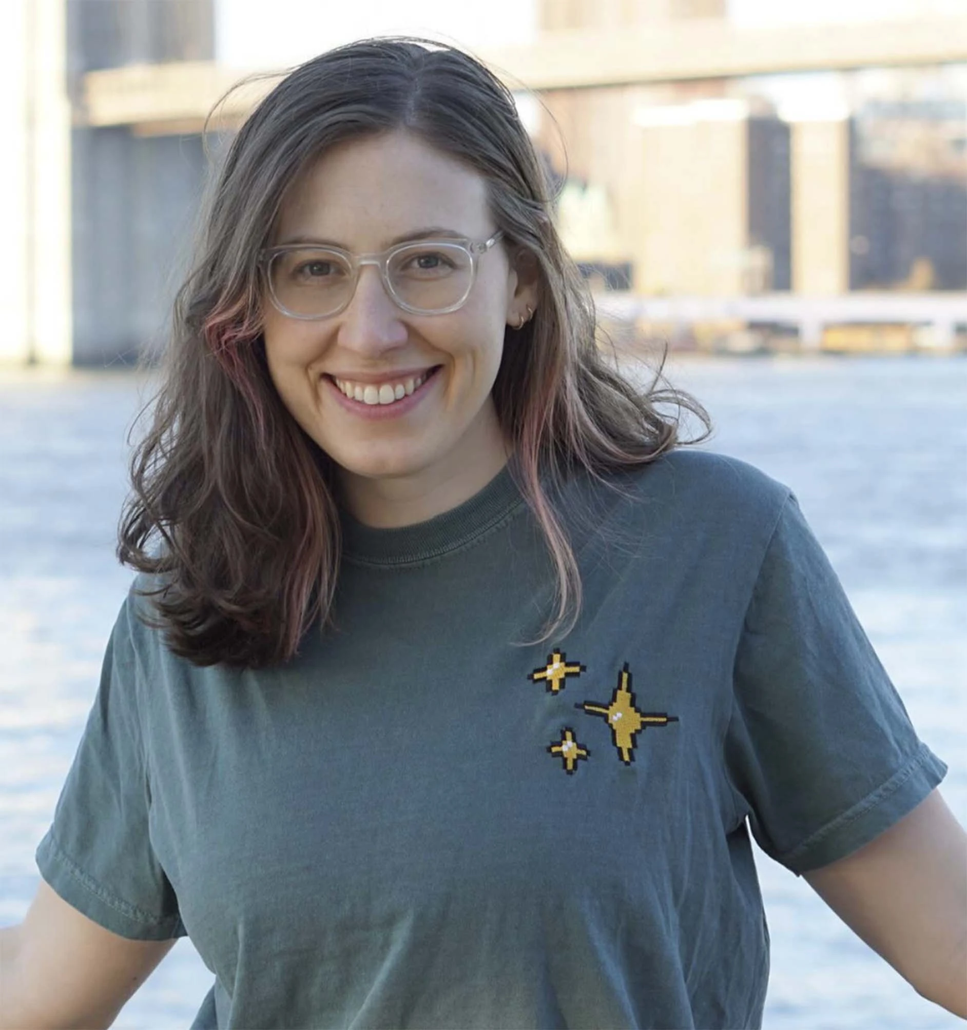 Femme souriante avec lunettes et t-shirt gris à motifs étoiles dorées, devant un pont et un plan d’eau.
