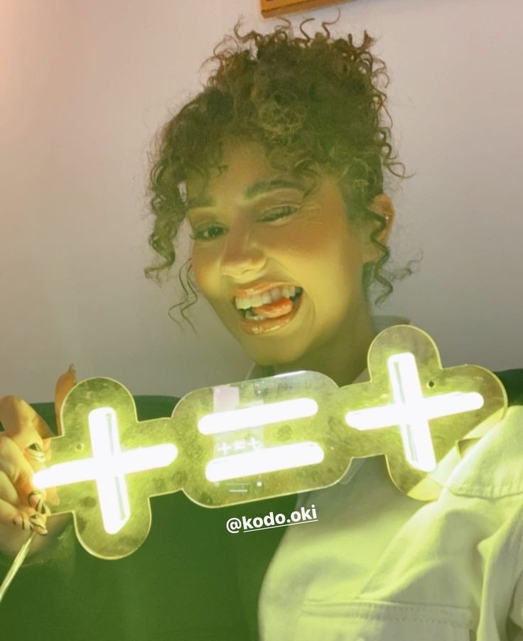 Femme souriante tenant un néon lumineux en forme de symbole ‘+=+’, ambiance fun et colorée.