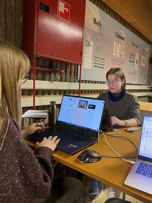 Deux personnes travaillant sur des ordinateurs portables dans un espace de coworking industriel, entourées de posters créatifs.